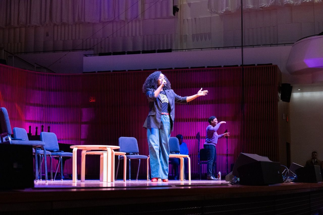 Woman with curly hair singing on stage with a microphone, another person in the background performing sign language, purple stage lighting, empty chairs and tables on stage.